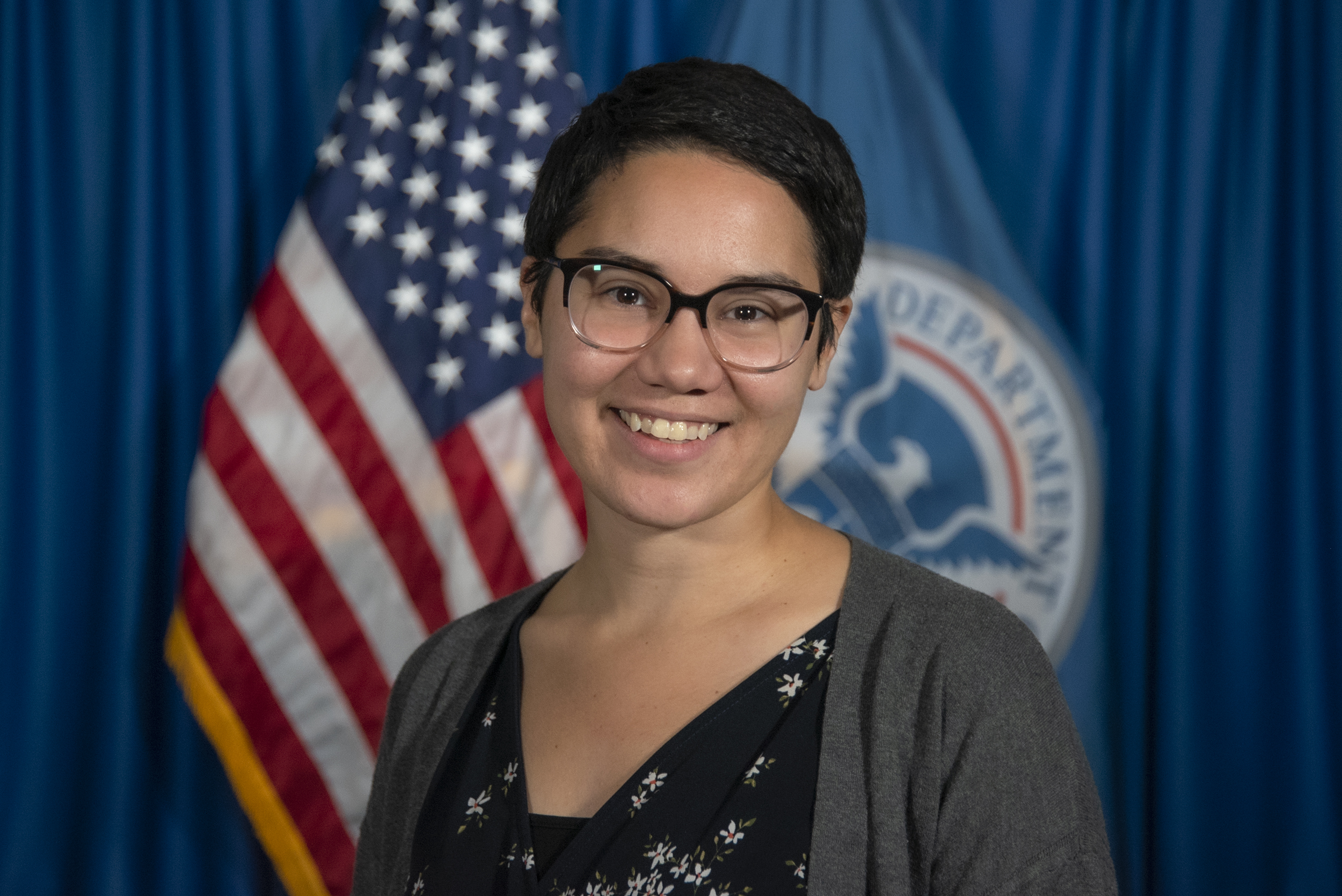 A photo of a woman in a gray cardigan and black shirt with short hair and glasses. in the background is the USA and DHS flag. 