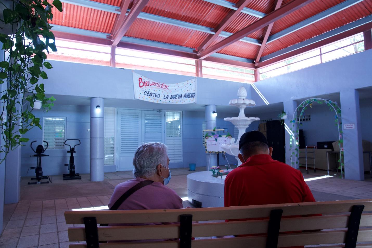 Two men sitting down on a bench in front os a fountain inside the CARMEN JOSEFINA “CAMBU” LÓPEZ RIJOS SENIOR CENTER. 