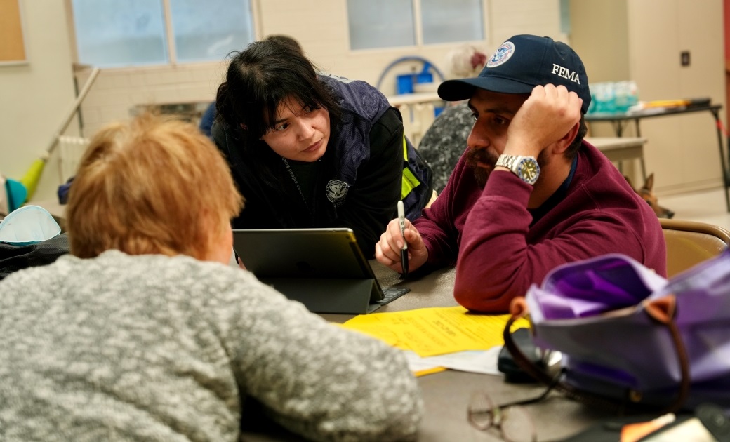 Two FEMA employees sitting with a disaster survivor and using a tablet.