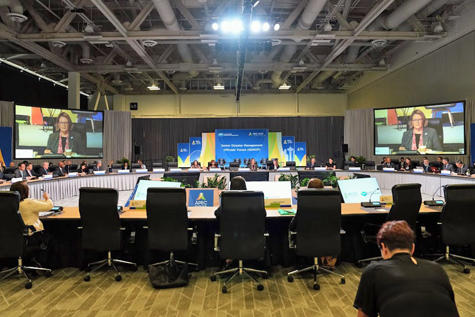 People in a conference room sitting at tables in a circle with speaker on large TVs.