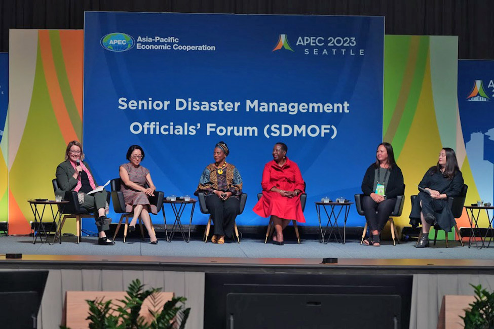 Six seated women sitting on a stage talking.