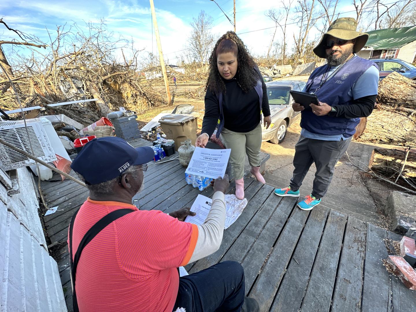Two FEMA employees hand a form to a man sitting on his porch.