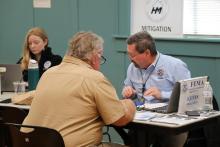 FEMA and Virginia Disaster Emergency Management staff members sitting at table across from survivors at a Disaster Recovery Center. Sign on the wall behind them reads "Mitigation."