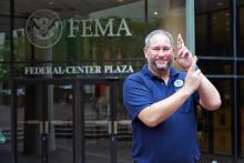 Aaron Kubey stands in front of a glass door at FEMA head quarters, wearing a blue polo shirt.