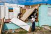 An elderly man stands in front of his damaged home while a woman is leaning over removing nails from a collapsed wooden wall 